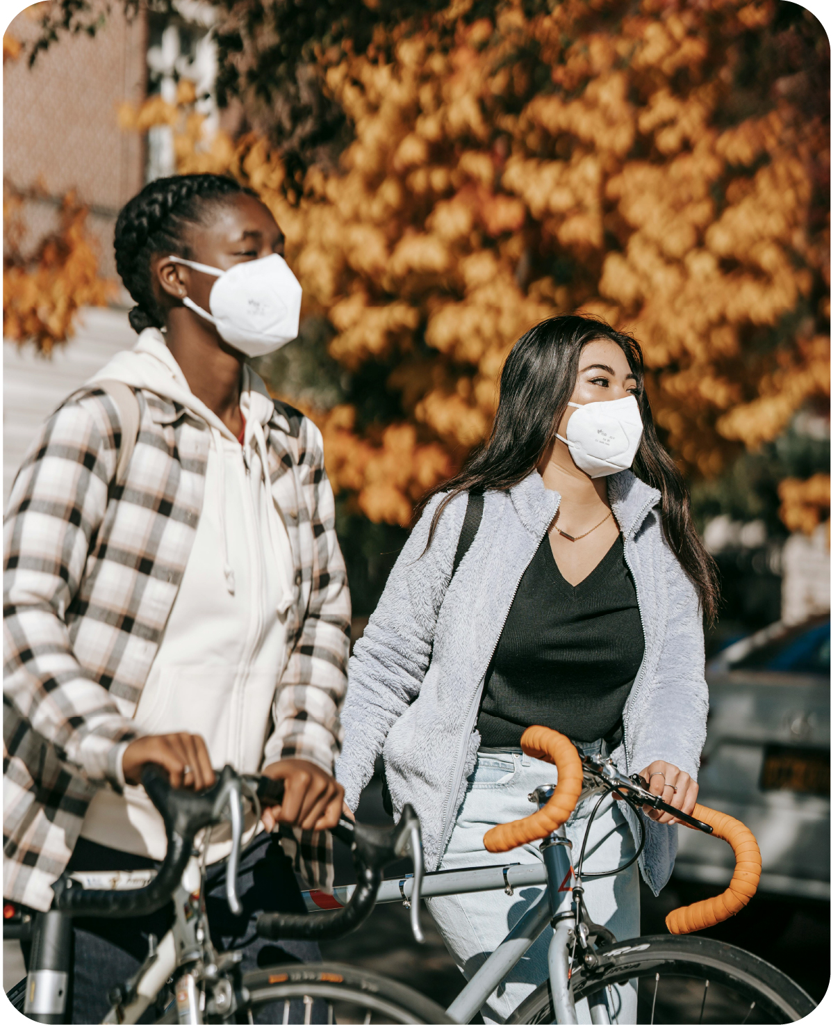 Two multiethnic friends in white kn95 masks walking their bikes near trees.