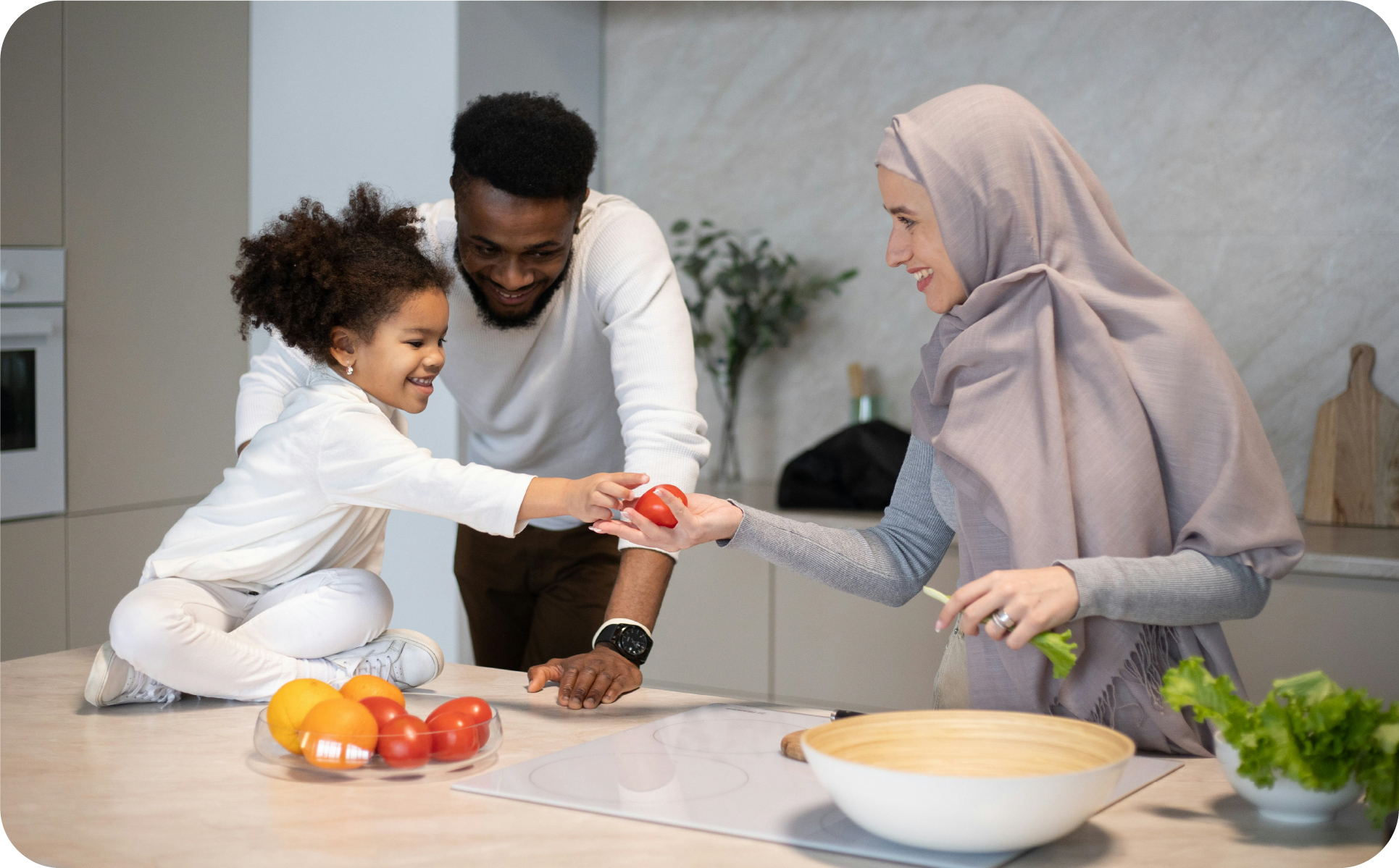 A mother in a hijab and a father in a kitchen, with their child sitting on the counter. They are all smiling and child is handing the mother a tomato. There is a bowl of tomatoes and a bowl of greens that the mother is handling.